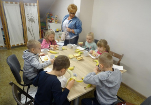 Sześcioro dzieci siedzi przy stole i jedzą ciasto, banana i popijają herbatą. Pani dyrektor stoi przy stole i spogląda na dzieci.